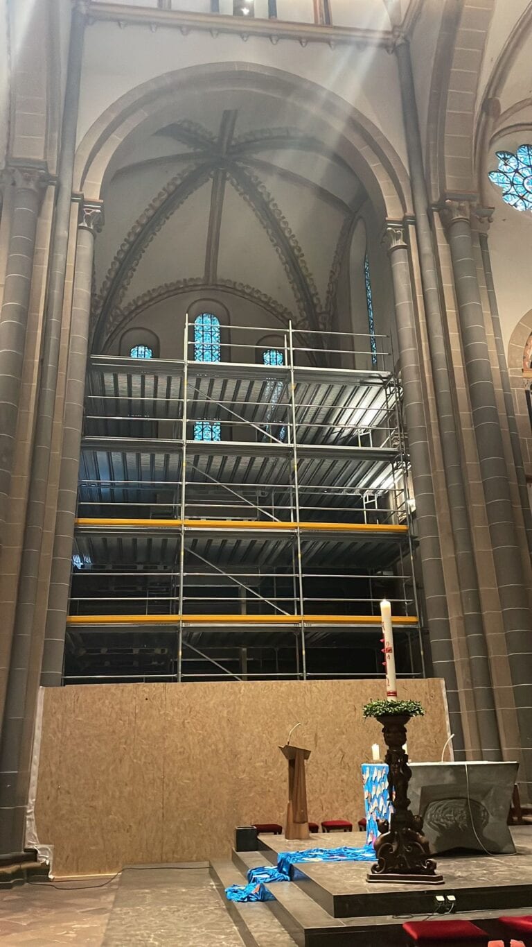 Innenraum einer Kirche mit Gerüsten und Kerzenleuchter, beige Wände, Buntglasfenster oben.