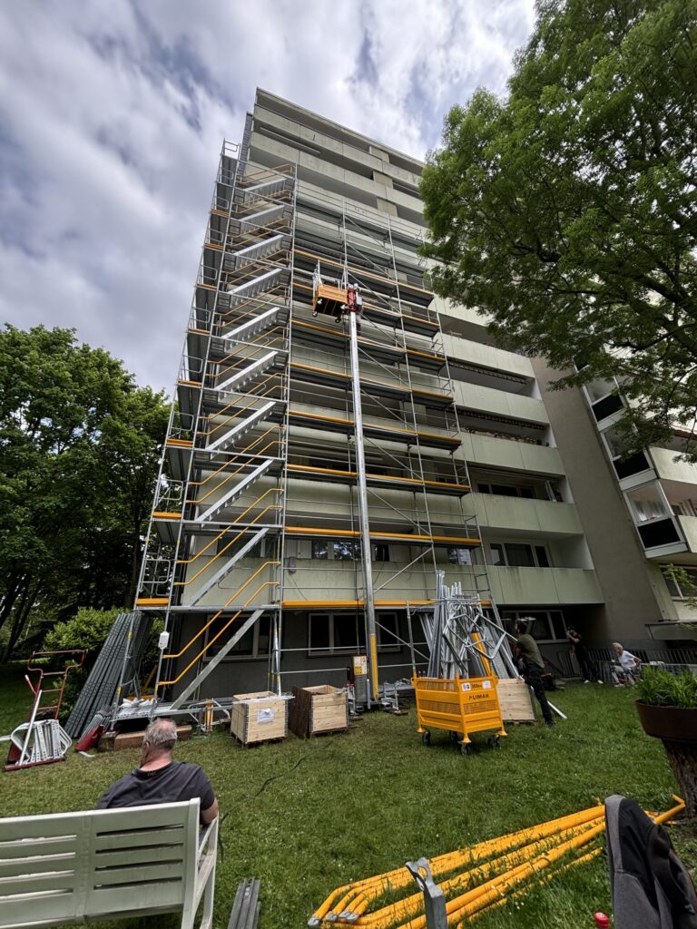 Gerüst an großem Gebäude, Person mit Helm arbeitet, grün-weiße Farben, Wolken am Himmel.