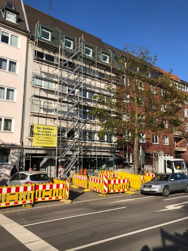 Ein Baugerüst an einem Wohngebäude, Straße gesperrt durch gelb-rote Absperrungen.